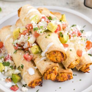 A stack of taquitos on a white plate with tomato and avocado on top for garnish.
