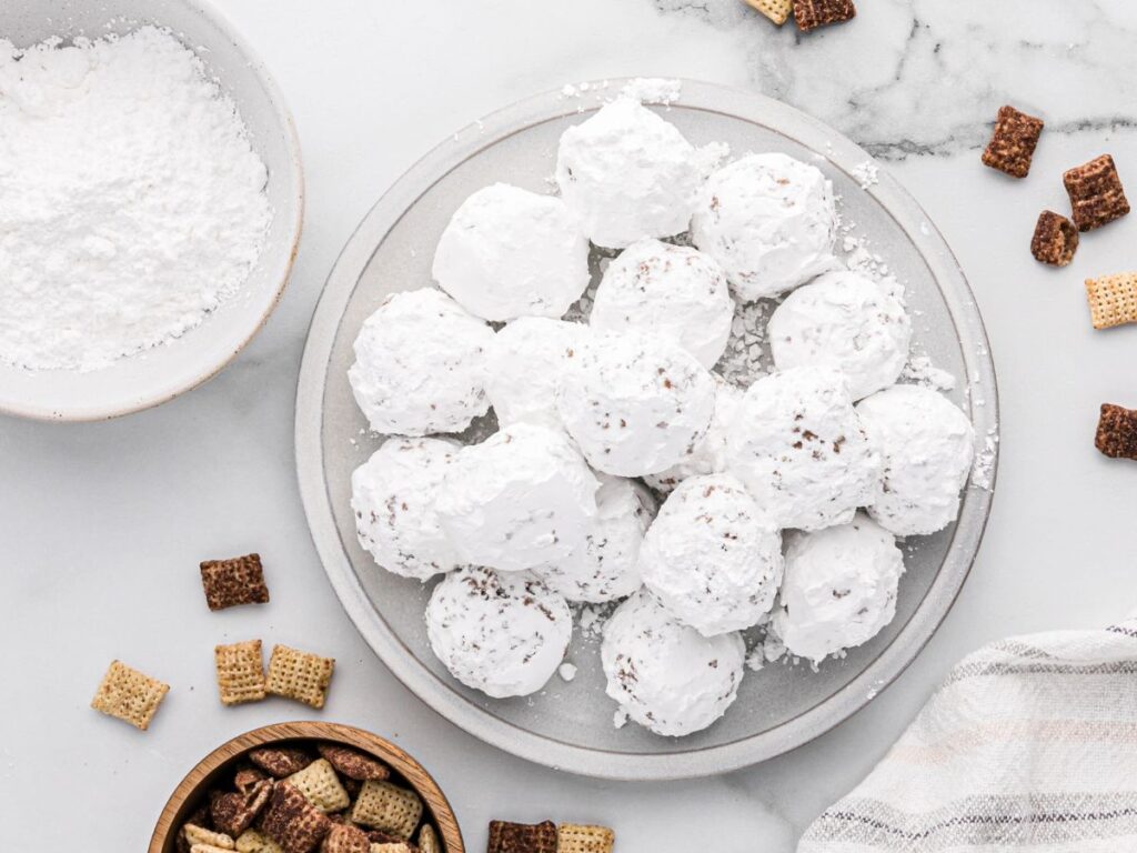 A plate of muddy buddy snack bites with a bowl of powdered sugar and chex cereal around the plate. 