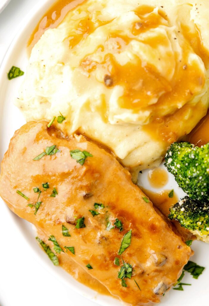 Serving of gravy pork chops on a plate with mashed potatoes and broccoli. 