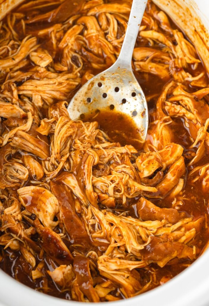Crockpot full of saucy shredded chicken with a stainless steel serving spoon in it. 
