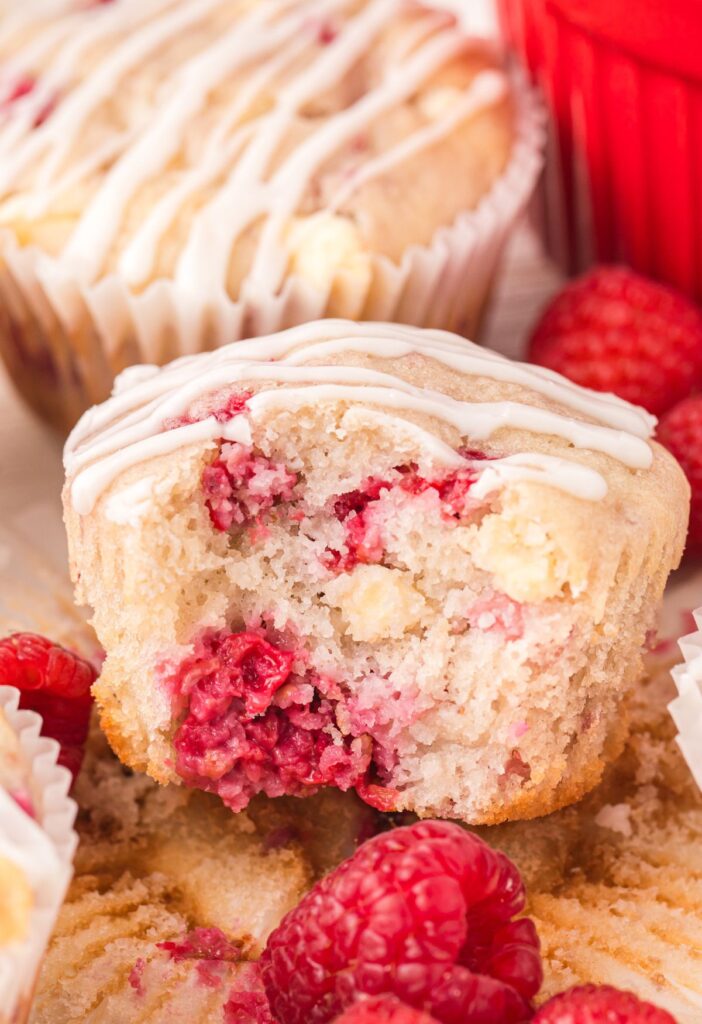 A muffin with a bite out of it against a red dish and fresh raspberries. 